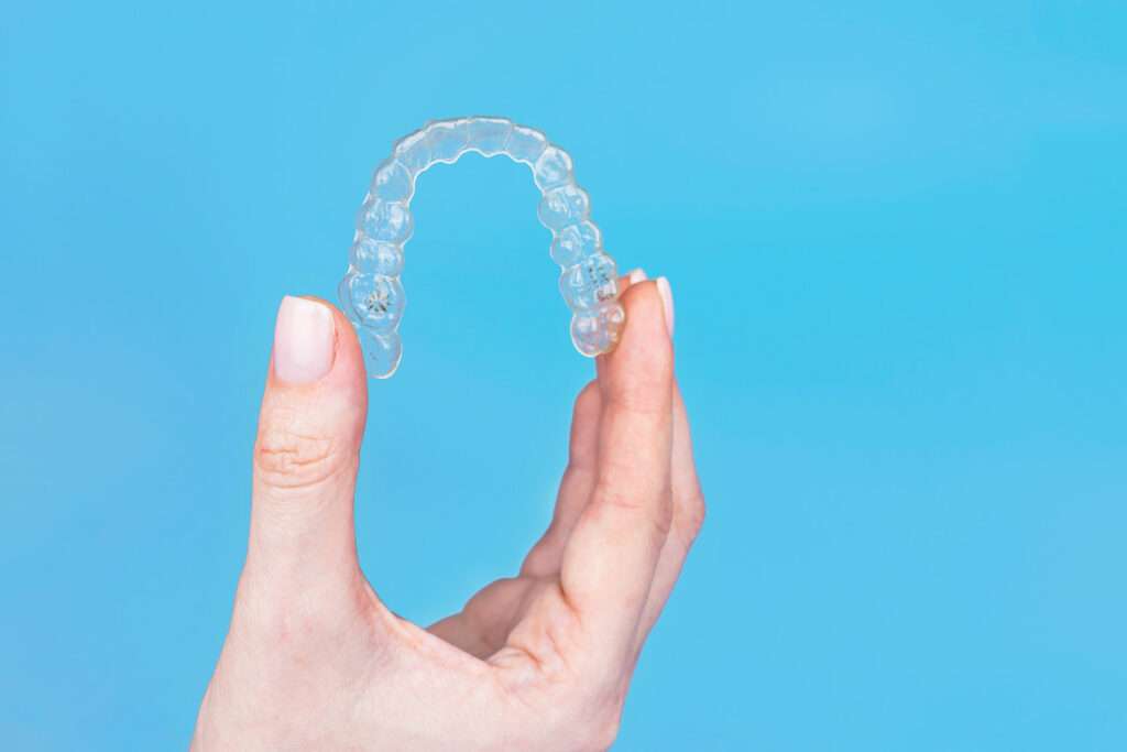 A woman with painted nails holds a clear aligner between her thumb and pointer finger.