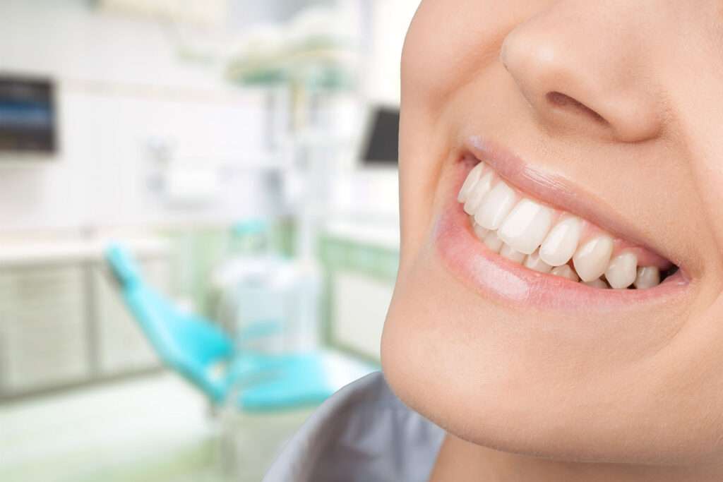 Woman smiling from the nose down in front of a dental examination chair.