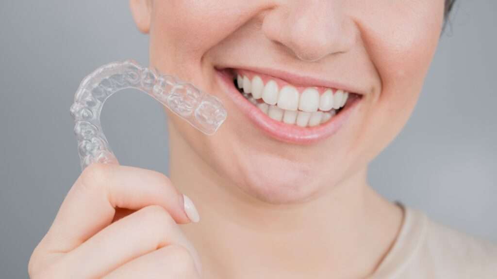 Woman with painted nails holding a clear aligner and smiling from the nose down to her shoulders.