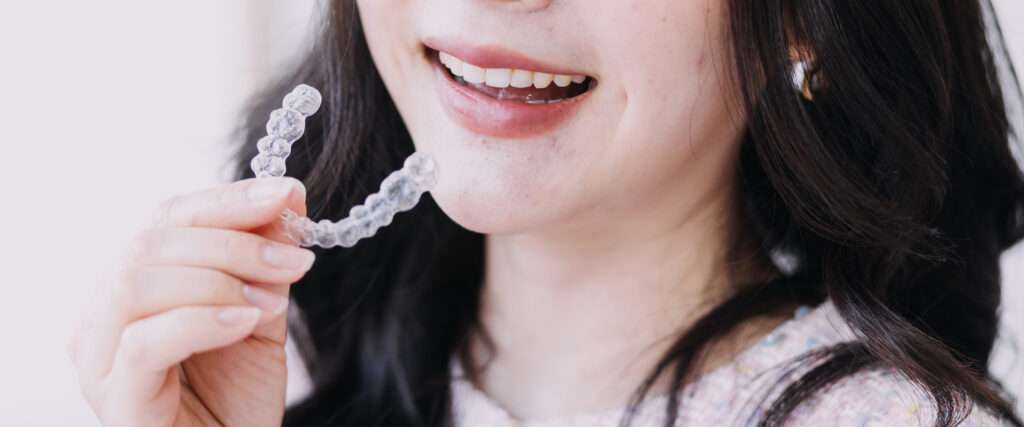 Woman with brown hair holding a clear aligner smiling from her lips down to her shoulders.