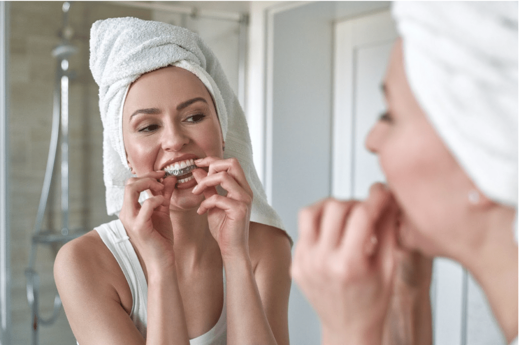 A young woman wearing a towel puts in her clear aligners, smiling at her reflection in the mirror.