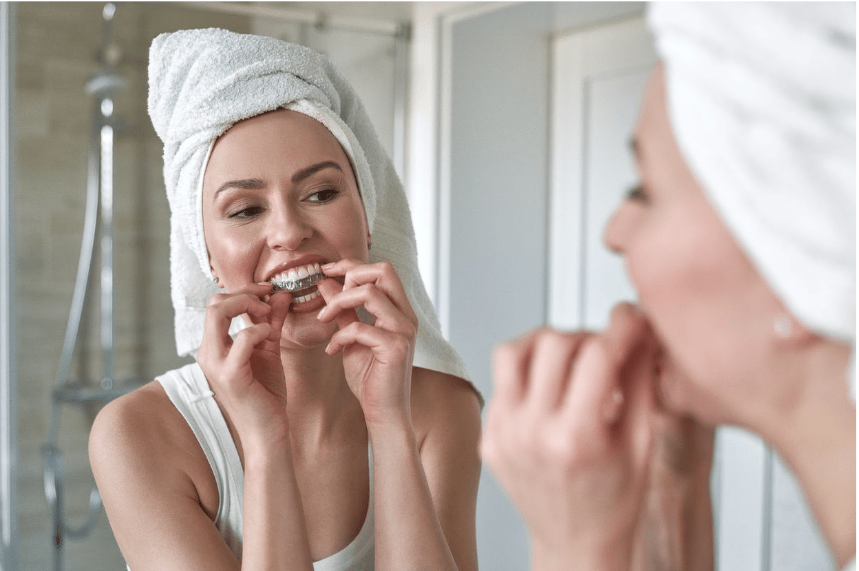 A young woman wearing a towel puts in her clear aligners, smiling at her reflection in the mirror.