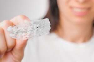 A woman holds up a pair of clear aligners. The aligners are in focus and she is blurry in the background.