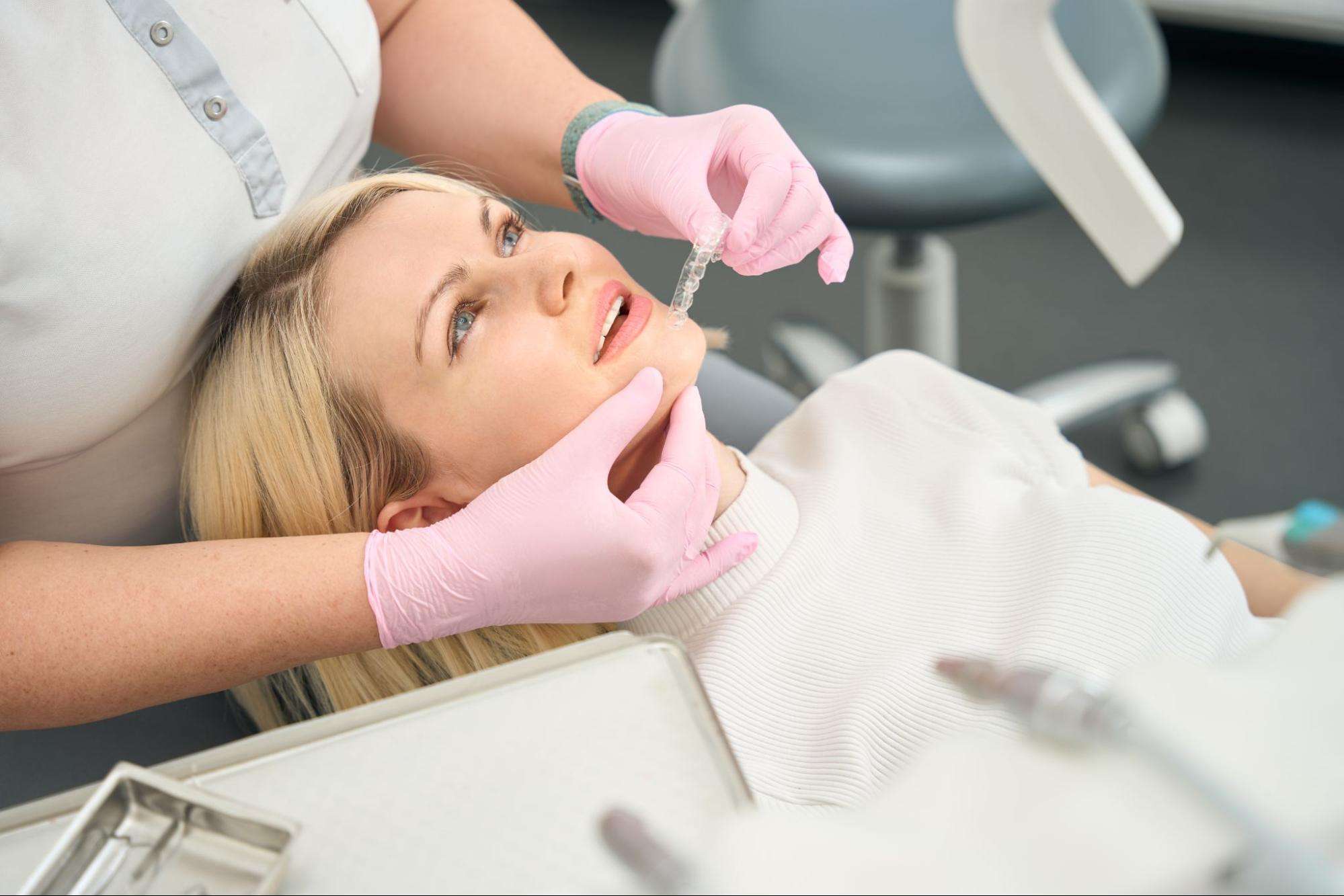 A young woman sits in a dentist’s chair while a staff member brings a clear aligner to her mouth.