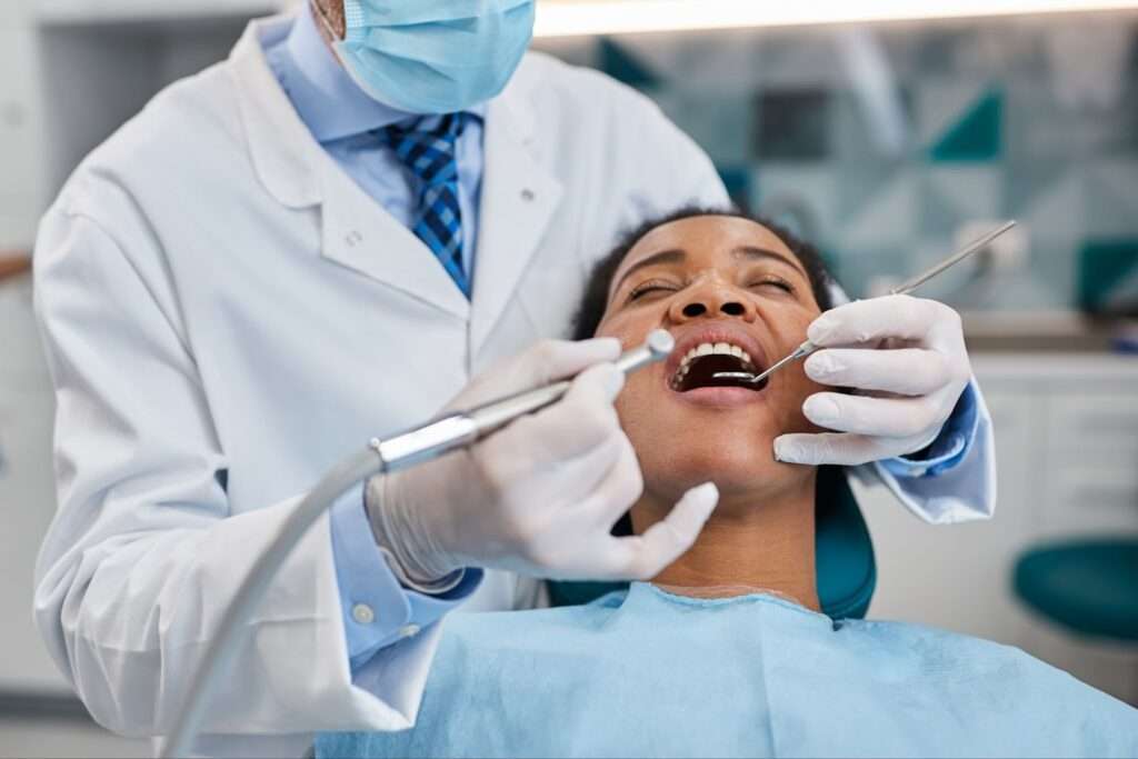 A dentist wearing gloves and a mask examines a patient's open mouth with dental tools in a clinic.