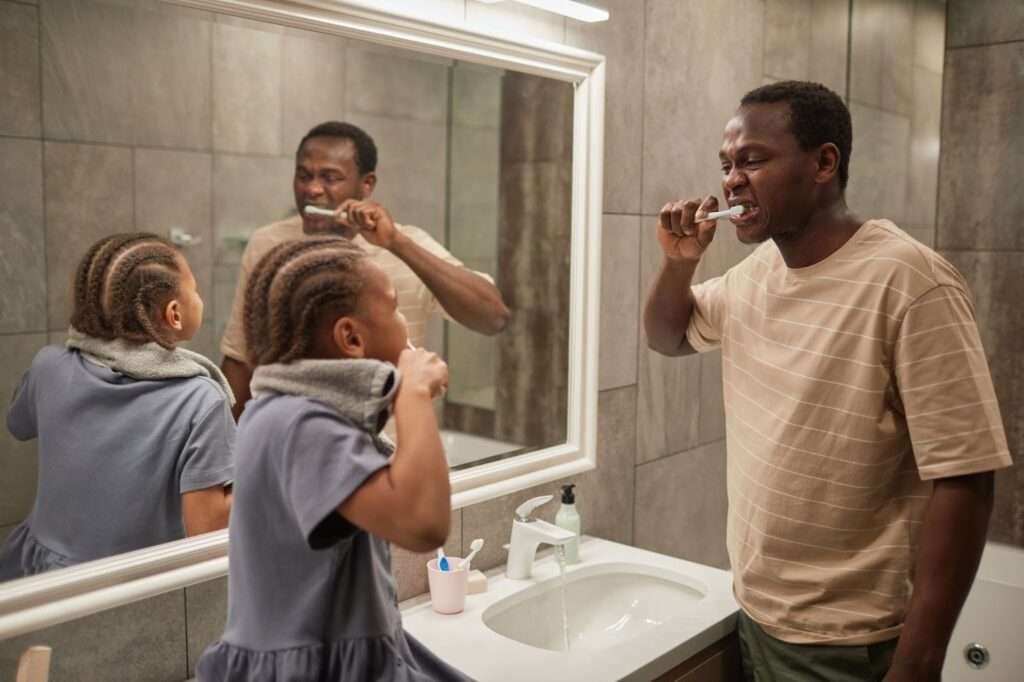 A man and a young girl stand in front of a bathroom mirror brushing their teeth. The girl has a towel on her shoulders and a toothbrush in a pink cup is visible on the sink.