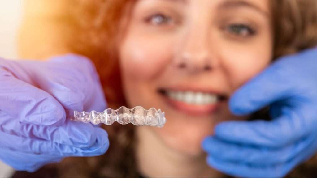 A close-up of a dentist's gloved hands about to apply an invisible aligner to a female patient's teeth during a dental appointment.