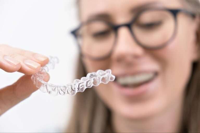 A smiling adult woman wearing dark-rimmed glasses holds a clear plastic aligner.