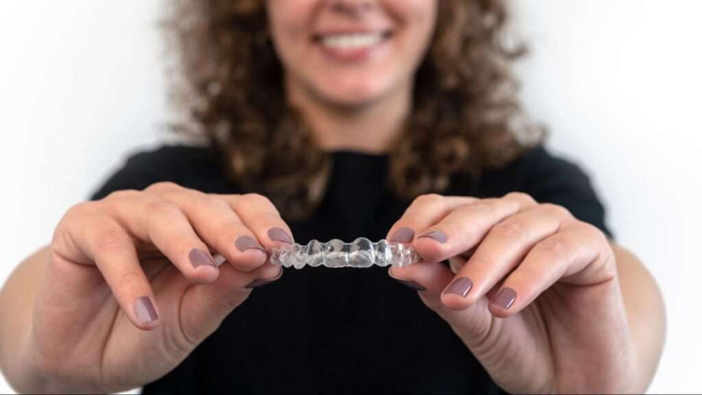 A cropped shot of a Caucasian woman with curly hair, wearing a black top, holding her invisible silicone aligner.